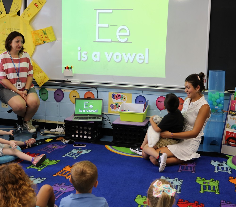 Teacher with student on her lap, while other students and teacher look at screen and learn the vowel E