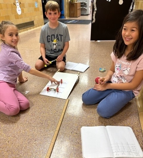 Three Wilson 3rd graders on floor with science project of designing and testing cars
