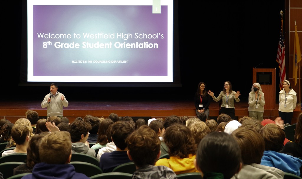 Edison principal speaks to 8th graders in auditorium with  screen behind him that says "Welcome to WHS's 8th Grade Student Orientation"