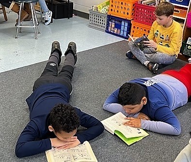 Two 4th grade boys stretch out on floor to read while another boy sits crosslegged while reading