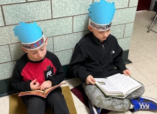 Two Jefferson students wearing handmade hats, sitting in hallway reading