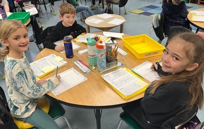 McKinley 2nd graders smile for camera as they sit at desk and work on writing assignment