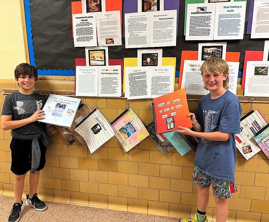 Two Wilson 4th graders hold their historical fiction project and smile for camera, in front of bulletin board with classmates projects